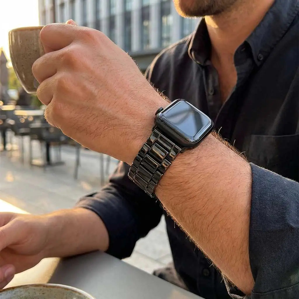 Anhem Classic Ceramic Apple Watch band in black featuring a polished 3-link design worn on a man's wrist at a cafe.