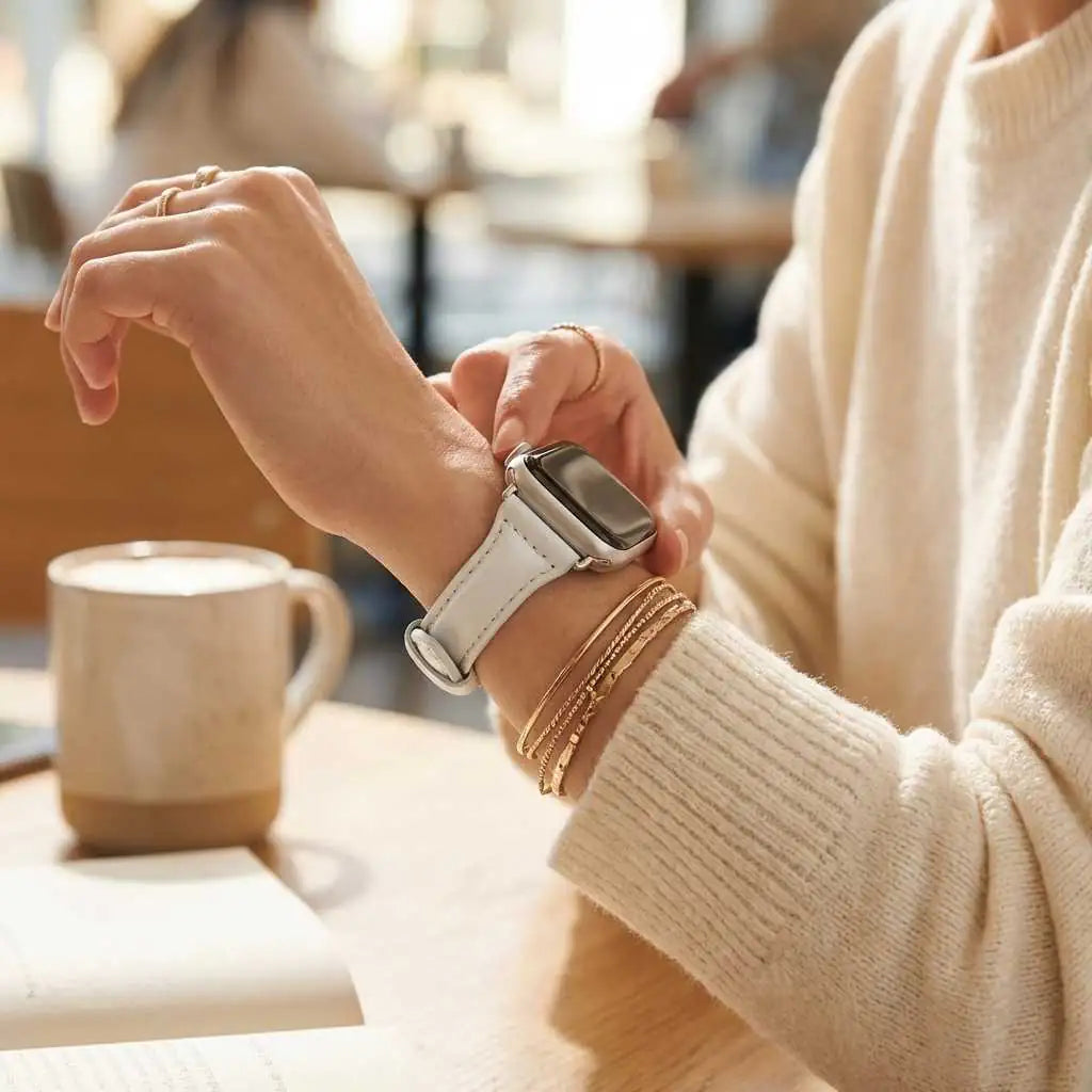 Anhem slim white leather Apple Watch band worn on a woman's wrist with a cream sweater in a cafe setting