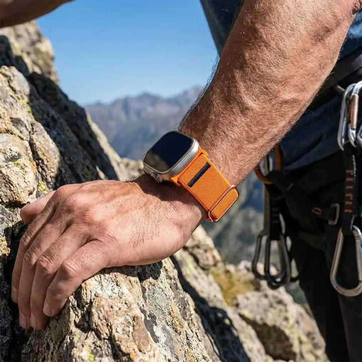 Anhem Ultra Nylon Strap in bright orange on a climber's wrist against a rocky mountain background.