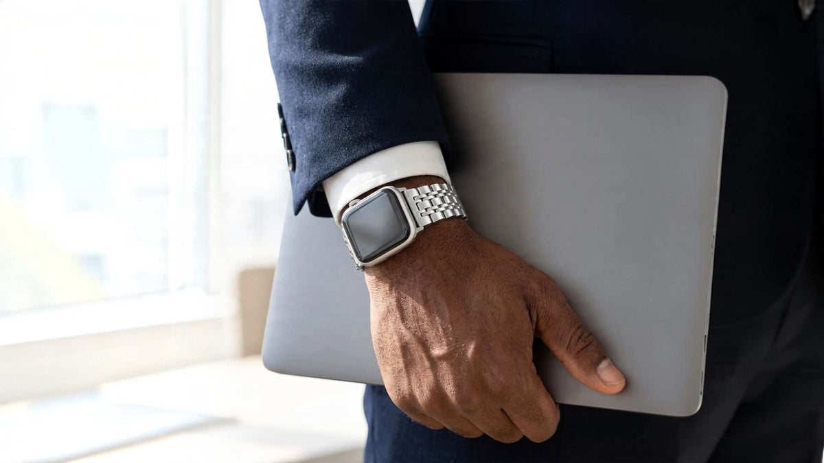 A close-up of a person in a dark navy suit holding a silver laptop under their arm, wearing a silver apple watch with a metal apple watch link band.