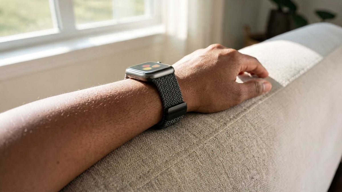 A close-up of a person's arm resting on a beige fabric couch, wearing a apple watch  with a black nylon band in a brightly lit room near a window.