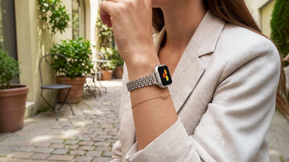 A woman in a light beige blazer wearing a silver apple watch band with a silver apple watch watch, standing in a sunlit outdoor patio setting.