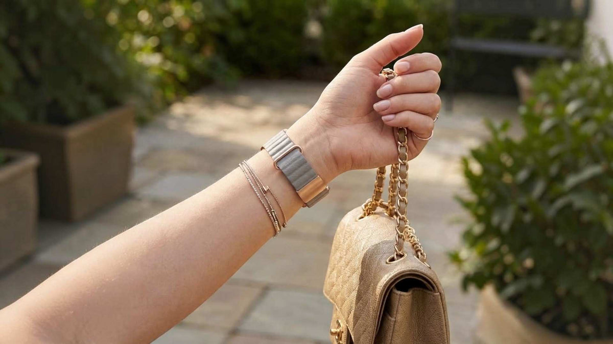 A close-up of a person's hand holding a beige handbag with a gold chain strap, wearing a rectangular silver-toned apple watch and delicate bracelets, set against a blurred outdoor garden background.