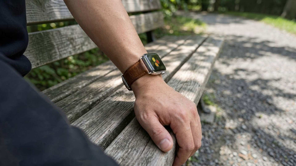 A close-up of a person's hand resting on a wooden park bench, wearing a apple watch with a brown leather strap and a silver-toned case in an outdoor setting.