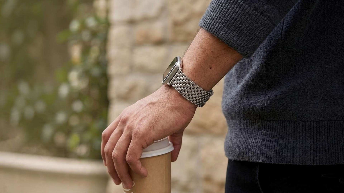 A close-up of a person's hand resting on a paper coffee cup, wearing a silver apple apple watch metal band with a dark rectangular face. The person is wearing a dark navy knit sweater, and the background is a softly blurred stone wall and greenery.