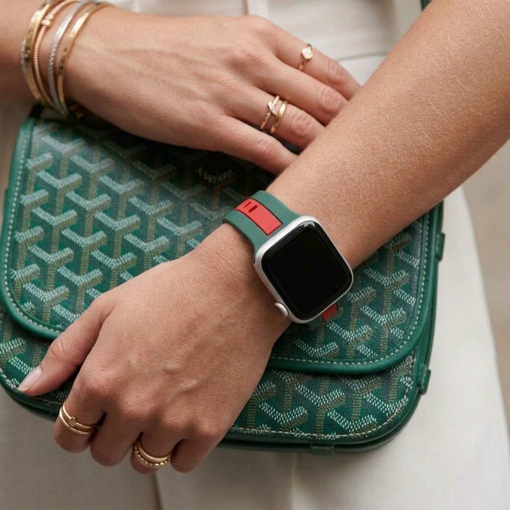 A close-up lifestyle shot of a person's hands and wrist wearing a green and red apple watch band with a silverapple watch . The person is holding a green patterned Goyard handbag.
