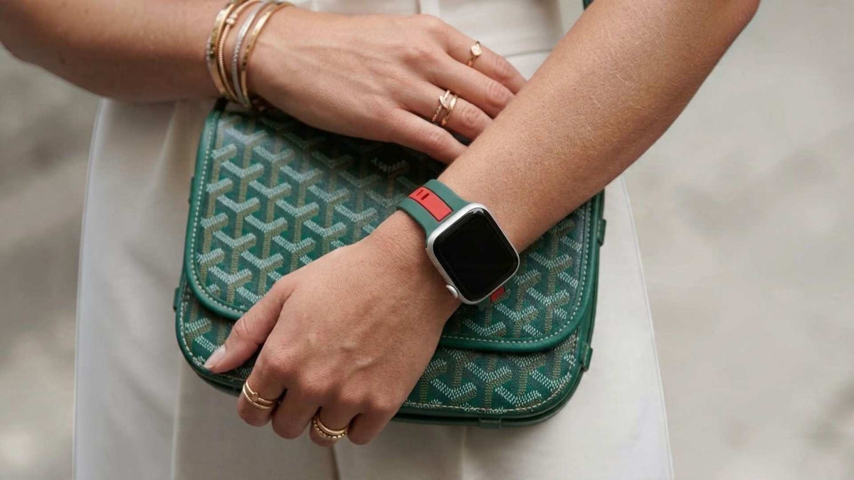 A close-up lifestyle shot of a person's hands and arms resting on a teal Goyardine handbag, wearing a apple watch with a green and red band.