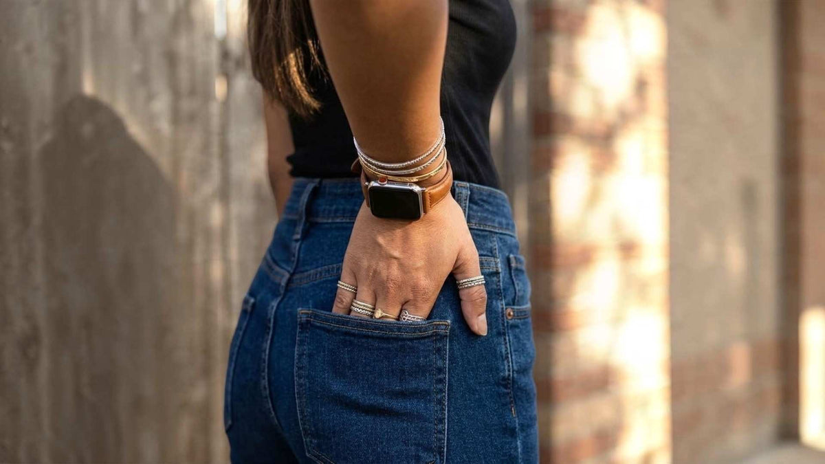 A close-up lifestyle shot of a person wearing a silver apple watch with a brown leather apple watch strap and stacked silver bracelets, paired with blue jeans, standing against a blurred brick wall.